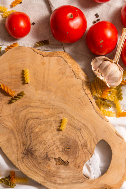 Auf der Tischdecke liegen bunte Nudeln, Tomaten und Knoblauch um ein Olivenbaum-Schneidebrett verstreut. Fragment. - Foto, Bild