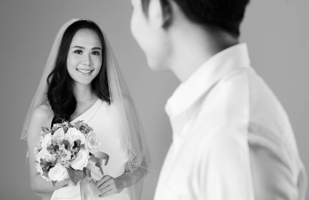Joven pareja asiática atractiva, enfoque selectivo en novia con vestido de novia con velo que sostiene ramo de flores. Hombre borroso con camisa blanca en primer plano. Blanco y negro. - Foto, imagen
