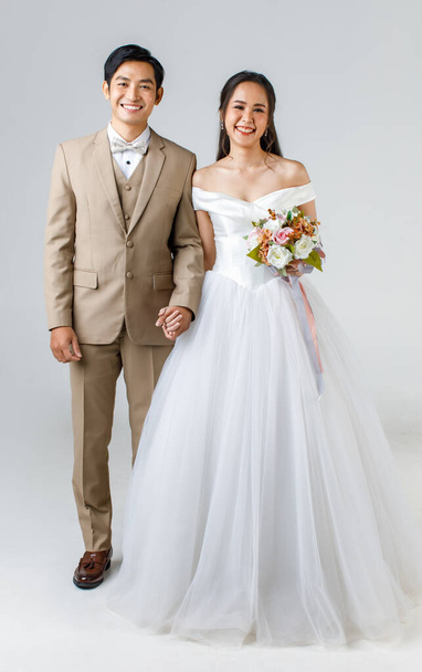 По всей длине молодая привлекательная азиатская пара, мужчина в бежевом костюме, женщина в белом свадебном платье, стоящая вместе, держась за руки. Концепция предсвадебной фотографии. - Фото, изображение