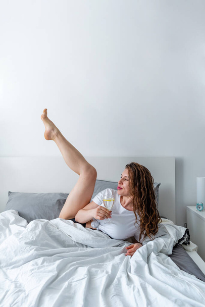 Young woman ginger hair doing yoga the bed in bedroom and drinks water with lemon. Fresh morning. Wellness and detox concept. Copy space - Fotografie, Obrázek