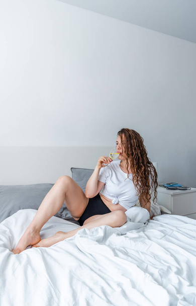Young woman ginger hair doing yoga the bed in bedroom and drinks water with lemon. Fresh morning. Wellness and detox concept. Copy space - Фото, зображення