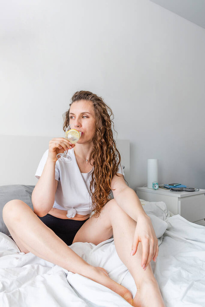 Young woman ginger hair sitting the bed in bedroom and drinks water with lemon. Fresh morning. Wellness and detox concept. Copy space - Foto, imagen