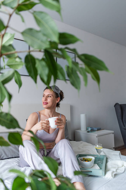 Young woman in silk pajamas has healthy breakfast while lying on bed in bedroom. Home wellbeing concept. Emotional health of a young woman - Valokuva, kuva