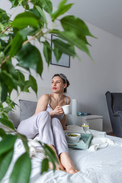 絹のパジャマの若い女性は寝室に寝そべっている間健康な朝食を有する。家庭の幸福の概念。若い女性の感情的な健康 - 写真・画像
