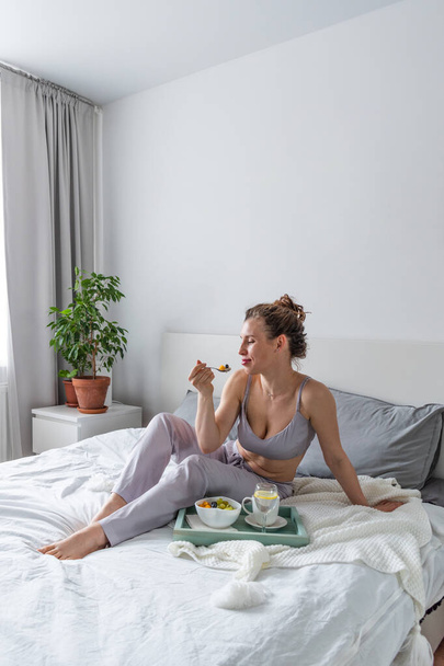 Young woman in silk pajamas has healthy breakfast while lying on bed in bedroom. Home wellbeing concept. Emotional health of a young woman - Foto, imagen