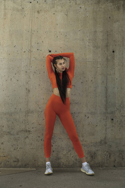Kobieta w pomarańczowej odzieży sportowej odpoczywa, by rozciągnąć triceps. Kobieta stojąca ubrania treningowe. - Zdjęcie, obraz