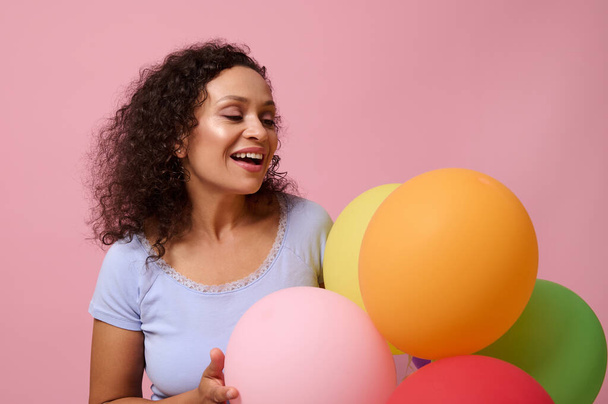 Beautiful mixed race with curly haired young woman rejoicing, posing against pink colored background with colorful air balls. Birthday party, anniversary, celebration, events concept with copy space . - Foto, afbeelding