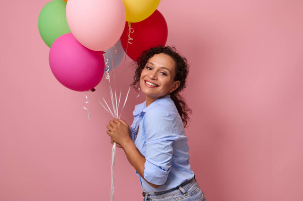 Ελκυστική όμορφη γυναίκα μικτής φυλετικής εθνικότητας που κρατά πολλά πολύχρωμα φωτεινά αερόστατα που απομονώνονται σε ροζ φόντο με χώρο αντιγραφής για κείμενο και διαφήμιση - Φωτογραφία, εικόνα