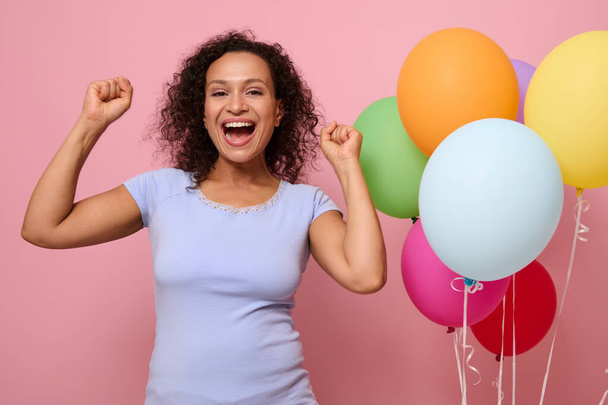 Studio shot of careless woman dances and has fun dressed in blue shirt and denim shorts, has festive mood, poses against pink wall background with gonflated balloons Concept of Time for having party. - Photo, image