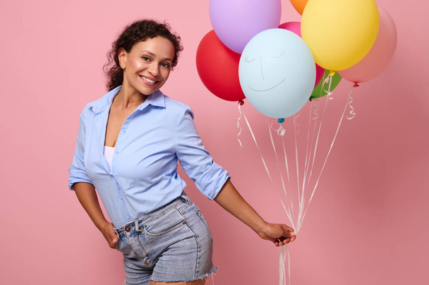 Cute young beautiful woman of mixed race ethnicity holds a lot of multicolored bright air balloons and looks at camera, isolated over pink background with copy space for text and advertising - Photo, image