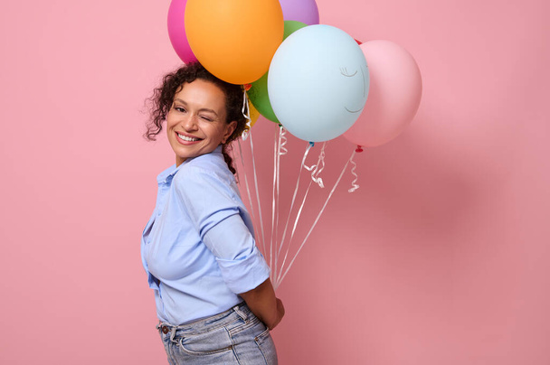 Приваблива приголомшлива жінка в блакитній сорочці посміхається з зубною посмішкою, дивлячись на камеру, тримаючи різнокольорові повітряні кульки за спиною, стоячи три чверті на рожевому кольоровому фоні, копіюючи простір
 - Фото, зображення