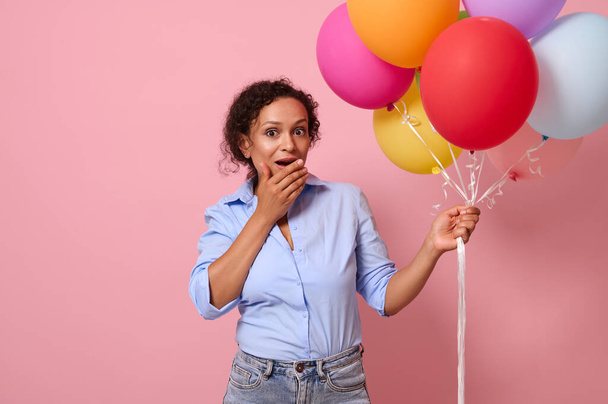 Sorprendido hermosa mujer afroamericana o hispana con globos de aire de colores en la mano cubre su boca, expresando asombro sorpresa. Aislado sobre fondo rosa con espacio de copia - Foto, imagen