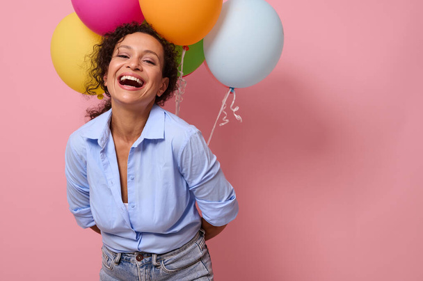 Iloinen nuori latinalaisamerikkalainen nainen iloisine ja iloisine tunteineen nauraa katsoen kameraan, pitäen ilmapalloja selkänsä takana, eristettynä vaaleanpunaiselle taustalle, jossa on kopiotilaa mainontaan - Valokuva, kuva