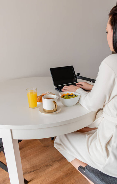 Young woman in comfortable white robe works on laptop while sitting at table the kitchen. Work at home for breakfast. Business woman maintains social distance, works remotely. New normal concept - 写真・画像