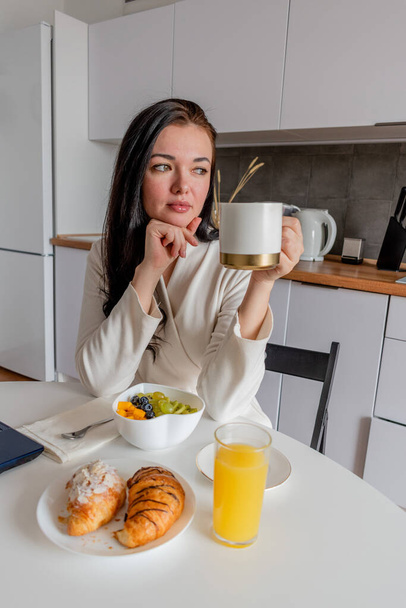 Mladá žena s černými vlasy v bílém županu sedí na židli v kuchyni. Ruce drží bílý hrnek s aromatickou kávou. Wellness a zdravá výživa koncept - Fotografie, Obrázek