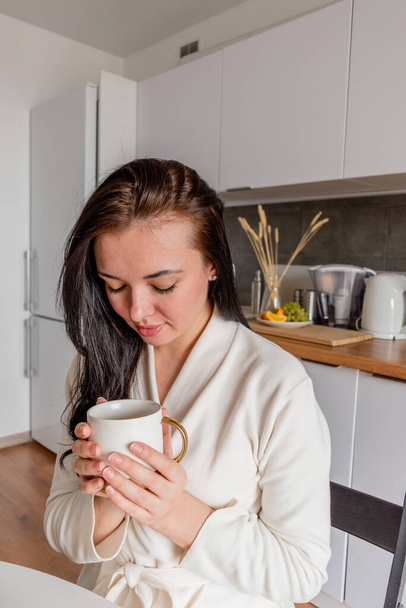 Young woman with black hair in white robe sits on chair the kitchen. Hands hold white mug with aromatic coffee. Wellness and healthy food concept - Фото, зображення