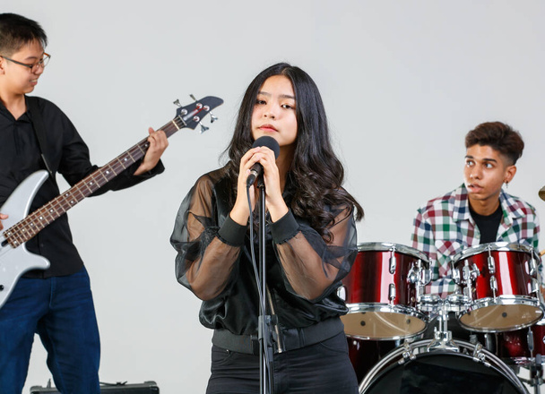 Group shot of teenage friends playing the music and singing together. Junior students play electric guitar, drum, and keyboard. A young female singer singing song with a musician in background - Foto, imagen