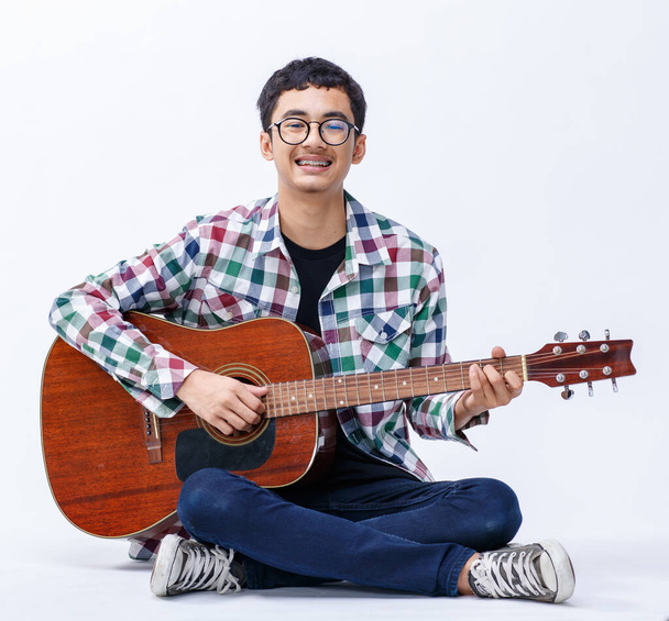 Retrato de un joven adolescente joven y sonriente sosteniendo la guitarra acústica. Guitarrista junior profesional sentado y tocando un instrumento mientras mira el fondo blanco aislado de la cámara - Foto, imagen
