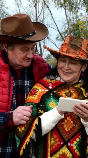 Felnőtt, középkorú, cowboy kalapos pár, akik futárral beszélgetnek, vagy nézegetnek valamit az okostelefonjukon. Az őszi parkban. Közelkép. - Felvétel, videó