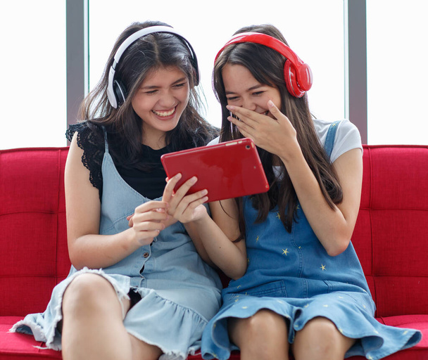 Zdjęcie portretowe młodych tajskich-tureckich nastolatków siedzących na czerwonej kanapie, oglądających filmy na czerwonym tablecie i głośno śmiejących się. Koncepcja mieszanej rasy sióstr mieszkających razem w domu - Zdjęcie, obraz