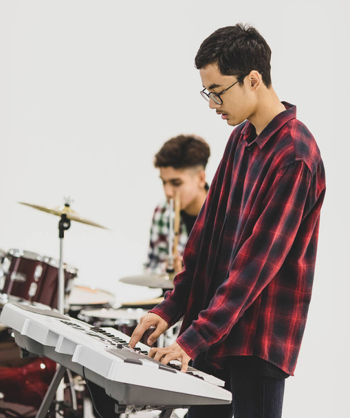 Portrait shot of teenage musician playing the keyboard. Selective focus at the Keyboardist with a drummer in the background. Professional junior student playing an instrument as a hobby with friends - Fotografie, Obrázek