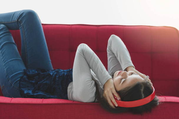 Retrato de un joven adolescente turco-tailandés tumbado en el sofá rojo y disfrutando escuchando música con auriculares rojos con las manos tocando el gesto de los auriculares. Concepto de hobby y relajación - Foto, Imagen