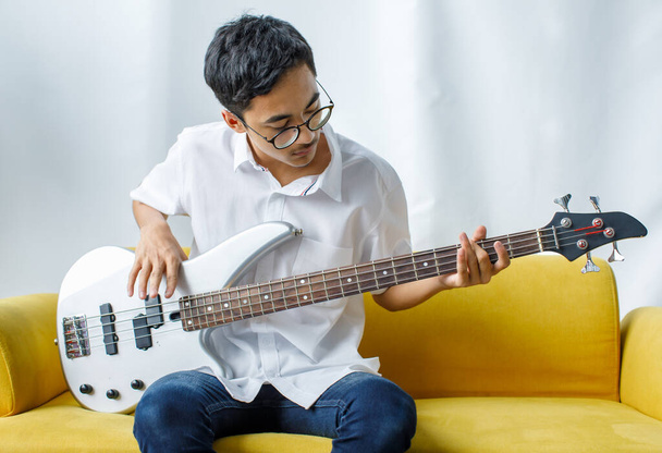 Portrét záběru hezkého usmívajícího se mladého teenagera, jak si užívá hraní na baskytaru. Junior kytarista v neformálním oblečení sedí na žlutém gauči a drží nástroj při pohledu do kamery - Fotografie, Obrázek