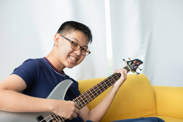 Portrait shot of handsome smiling young boy with eyeglasses enjoying playing the bass guitar. Junior bassist in casual clothes sitting on couch and holding instrument while looking at the camera - Photo, Image