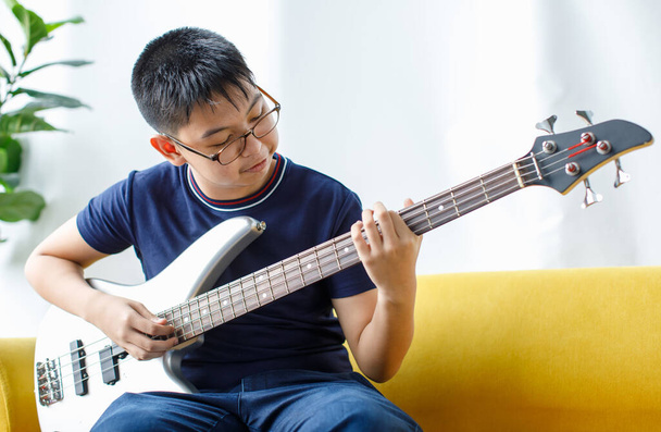 Портретний знімок гарного усміхненого молодого хлопчика з окулярами, який насолоджується грою на бас-гітарі. Молодий басист в повсякденному одязі сидить на дивані і тримає інструмент, дивлячись на камеру
 - Фото, зображення
