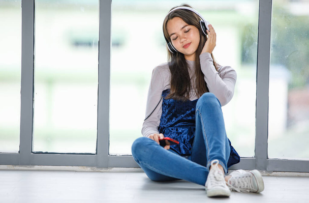 Portrét mladého thajsko-tureckého teenagera, jak poslouchá hudbu se sluchátky. Junior smíšené-závod dívka těší hudbu s rukama dotknout sluchátka a zavřené oko gesto při posezení na podlaze - Fotografie, Obrázek