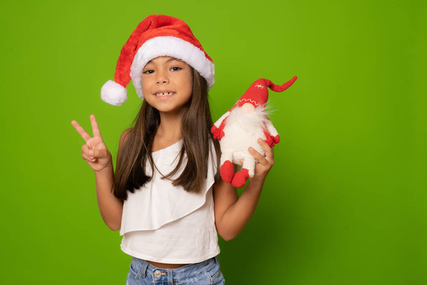 Glückliches kleines Mädchen mit Weihnachtsmütze und Siegeszeichen-Geste vor grünem Hintergrund. - Foto, Bild