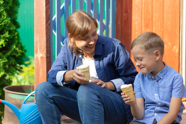 Óvodás fiú anyával, aki jégkrémet eszik egy ház verandáján a faluban egy nyári napsütéses napon. Szelektív fókusz. Portré - Fotó, kép