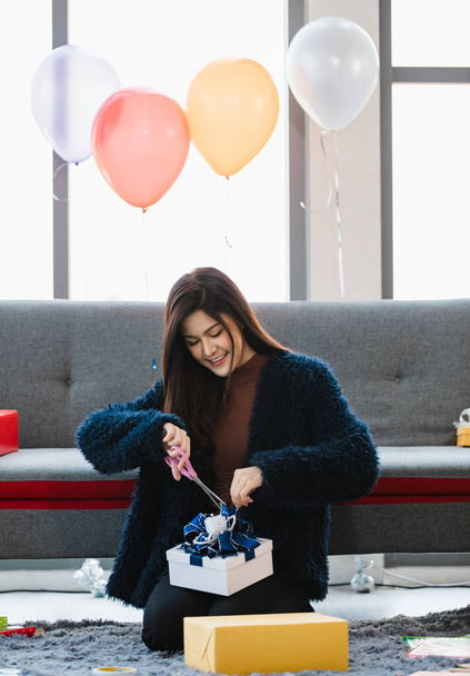 Ázsiai fiatal boldog csinos születésnapi lány szőrös kabát ül elöl szürke kanapén dobozok és játékmaci babák a nappaliban mosolygós használat olló vágás kék szalag fehér ajándék doboz. - Fotó, kép