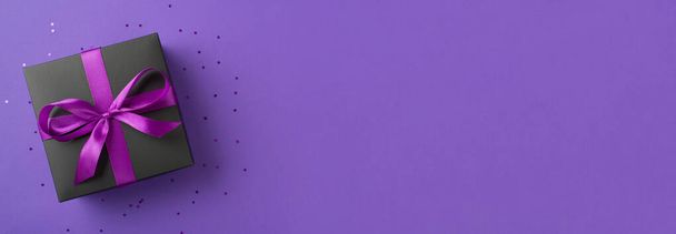 Foto vista superior de la elegante caja de regalo negra con lazo de cinta de satén púrpura y confeti sobre fondo violeta aislado con espacio vacío - Foto, imagen