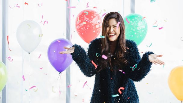 Aziatisch jong gelukkig mooi vrouw in blauw harige jas staande glimlachende blik op camera onder roze papier popper confetti in de voorkant kleurrijke Nieuwjaar feest viering decoratie ballonnen achtergrond. - Foto, afbeelding