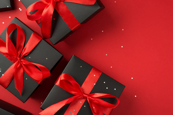 Фотографія подарункових коробок у чорній упаковці з бантом з червоною стрічкою та блискітками на ізольованому червоному тлі з копійкою
 - Фото, зображення