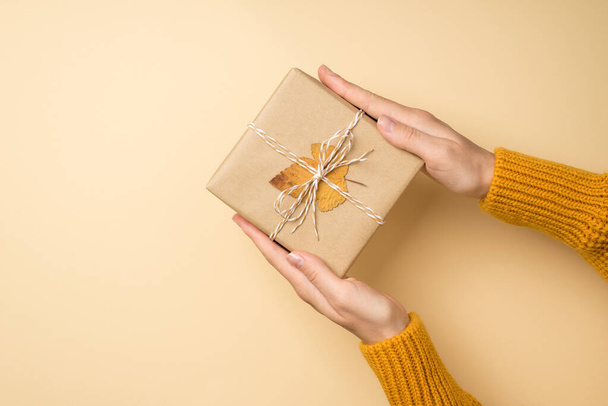 Πρώτη πρόσωπο top view φωτογραφία των χεριών σε κίτρινο πουλόβερ που κατέχουν craft χάρτινο δώροbox με φιόγκο νήματος και κίτρινο φύλλο φθινόπωρο σε απομονωμένο μπεζ φόντο με copyspace - Φωτογραφία, εικόνα