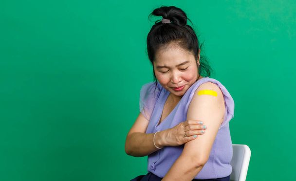 Studio shot of Asian middle aged woman sit smile and show yellow yaster bandage on arm after receive coronavirus covid 19 vaccination by doctor at clinic on green background. - Foto, imagen