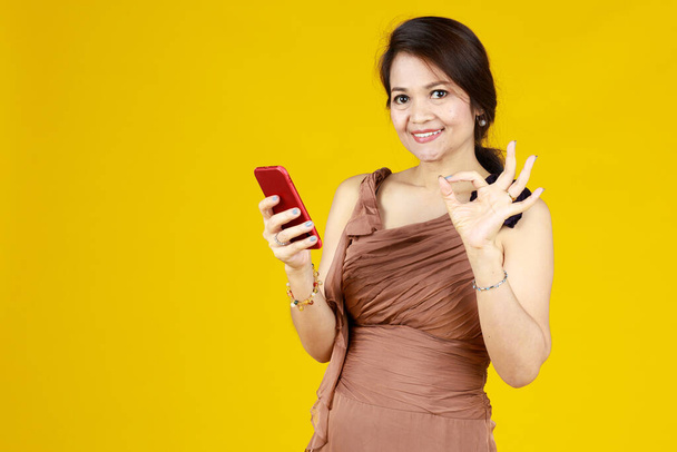 Krásná asijská dáma v hnědých šatech stojí s ok povzdechem. Druhá ruka drží červený mobil. Zdá se úspěšné získat slevu on-line nakupování jako její tvář ukazuje velmi šťastný - Fotografie, Obrázek