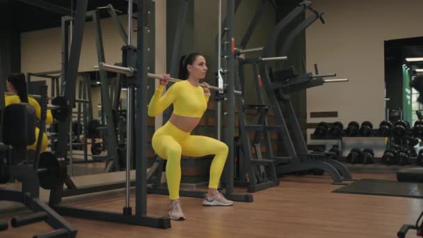 Edzés Spanyol fiatal nő testépítő izmok segítségével Inflight Fitness Smith Machine tornaterem vagy fitness klub. Lány emelje súlyát súlyzóval a vállán, miközben guggol testmozgás a jó egészség érdekében. - Felvétel, videó