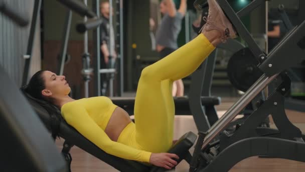 Hispánská žena pomocí tiskového stroje ve fitness klubu. Mladá žena cvičí na nohou v simulátoru ve žlutém sportovním oblečení, - Záběry, video