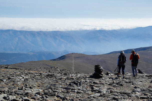 dva turisté kráčející po břidlicové zemi sestupující z vrcholu Mulhacenu, pak pláň křižovaná stezkou, pak pohoří a v pozadí moře nízkých mraků - Fotografie, Obrázek