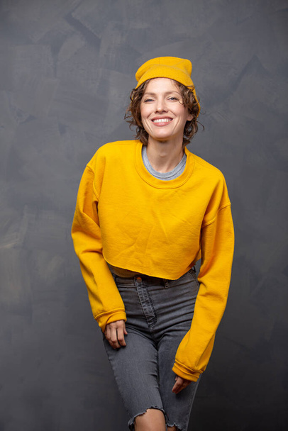 Autumn mood! Portrait of happy smiling woman with short brunette curly hair wearing yellow sweater and a hat over grey wall background - Valokuva, kuva
