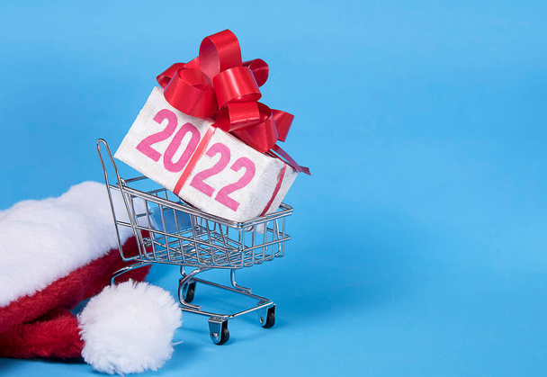 2022 text on gift box with red ribborn, santa claus hat and shopping trolley. Blue background. Christmas and New Year sale and shopping concept. Copyspace. - Фото, изображение