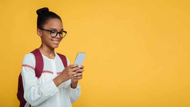 Vrolijke slimme jonge Afrikaans-Amerikaanse dame leerlingen in glazen met rugzak typen bericht op smartphone - Foto, afbeelding