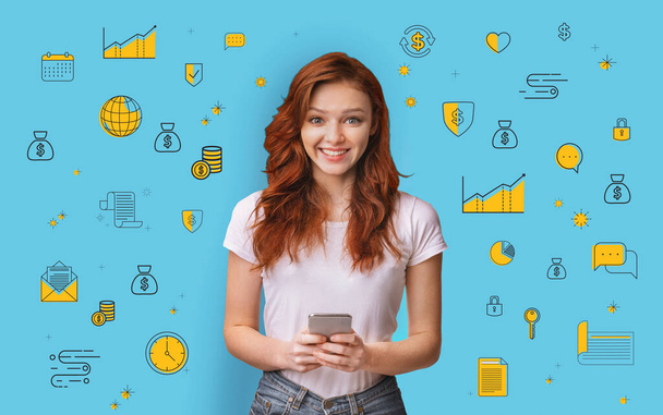 Happy millennial red haired woman using mobile banking application on blue studio background with money related icons - Photo, Image
