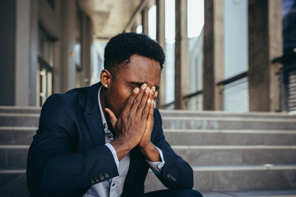 Man kantoormedewerker moe na het werk, Afro-Amerikaan heeft ernstige hoofdpijn, zittend op trappen in de buurt van kantoor - Foto, afbeelding
