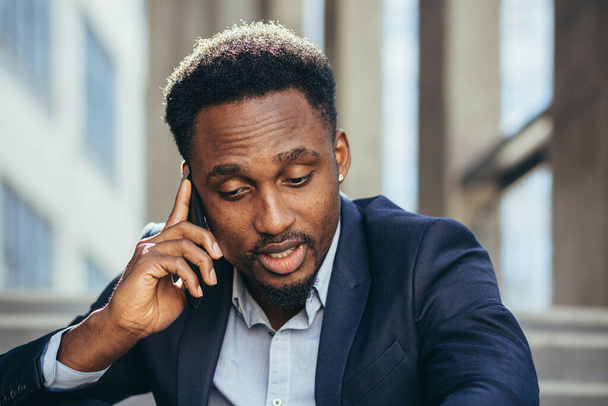 Depressed african american businessman telling sad news talking on cell phone sitting on office stairs in business suit - Photo, image