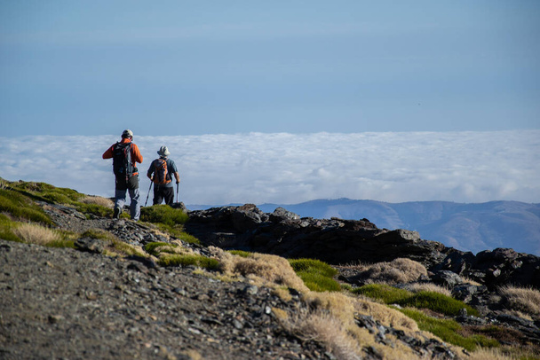 twee wandelaars wandelen door Sierra Nevada met struiken en een zee van wolken op de achtergrond - Foto, afbeelding