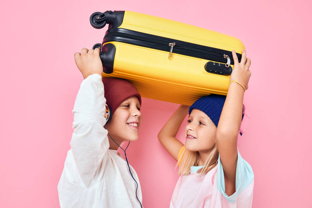 Portrait of a girl and a boy with a yellow suitcase in his hands pink color background - Foto, imagen